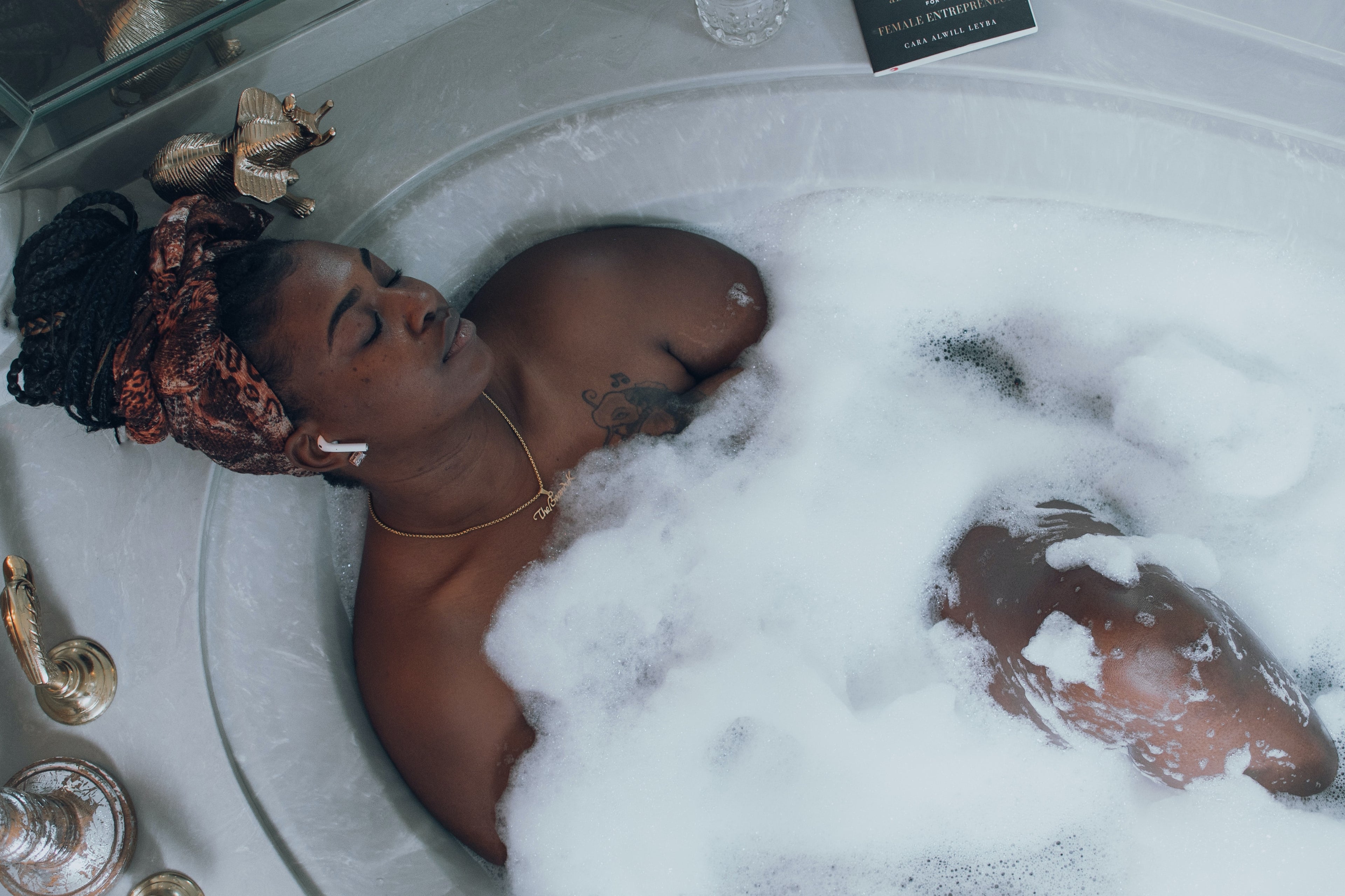 Brown skin woman soaking in bath tub surrounded by bubbles and listening to music in her portable headphones